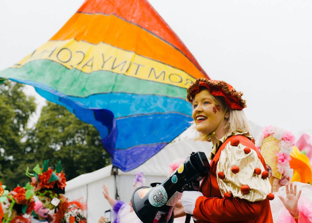 A woman in a red headress smiles and holds a megaphone in front of rainbow coloured flag